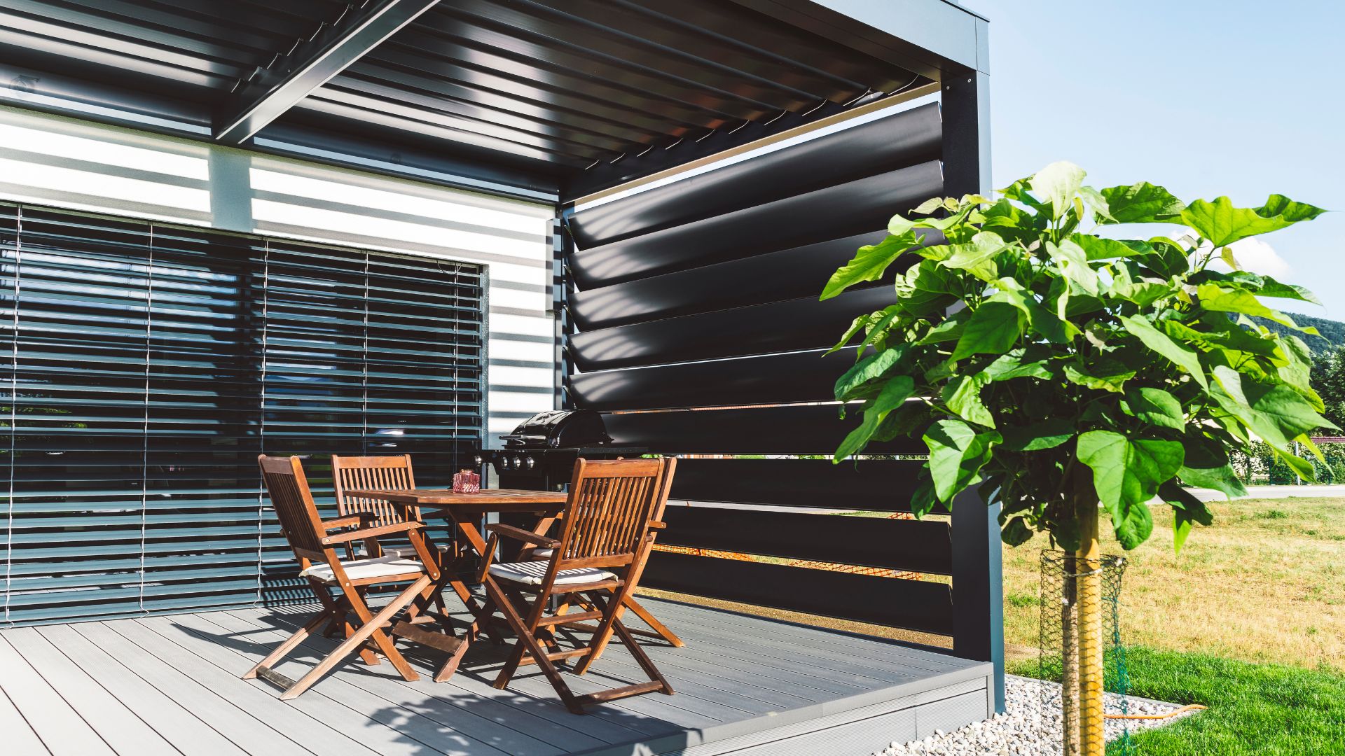 Contemporary black pergola with wooden table and chairs on a patio, featuring a large potted plant.
