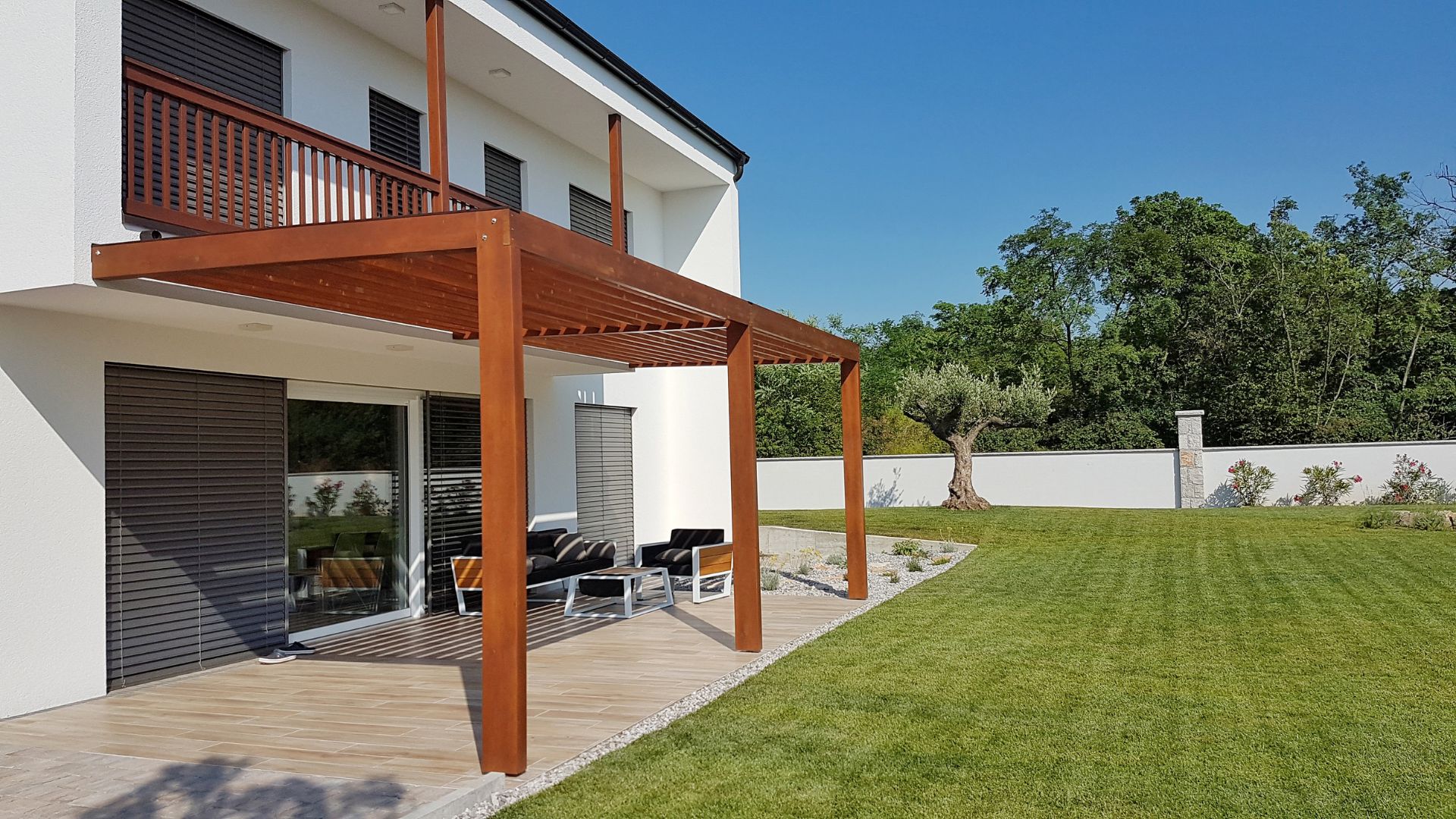 Wooden pergola extending from a white two-story house, providing shade over an outdoor seating area with a large green lawn.