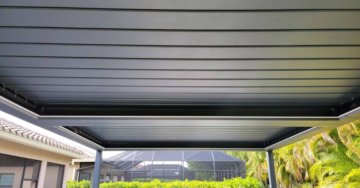 A close-up view of an all-black solid roof pergola ceiling with deep channels for water drainage, situated next to a residential home and pool cage.