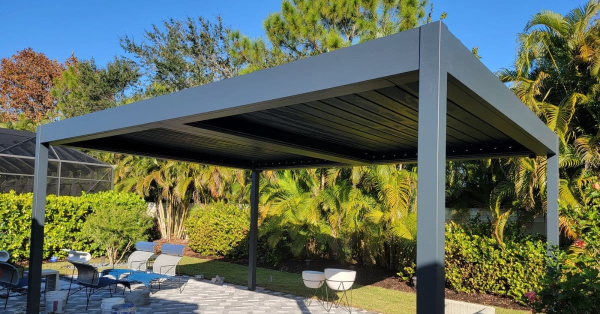A freestanding dark grey solid roof pergola installed on a grey stone paver patio, surrounded by tropical landscaping and outdoor lounge chairs.