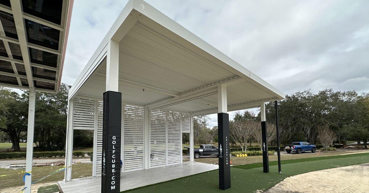 A white motorized pergola structure with adjustable louvers and slatted privacy walls, used as a professional golf hitting bay on a green turf area.
