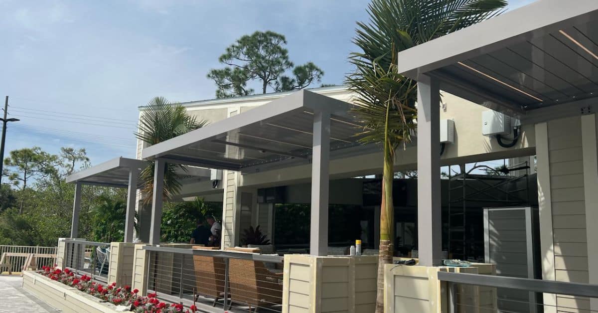 A grey residential motorized pergola system with integrated LED lighting installed over a luxury patio area with wicker seating and tropical landscaping.