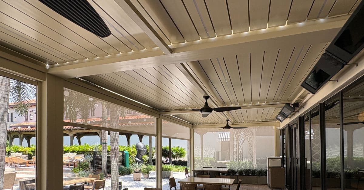 An interior view looking out from under a motorized louvered pergola with semi-transparent retractable privacy screens lowered to block wind and sun.
