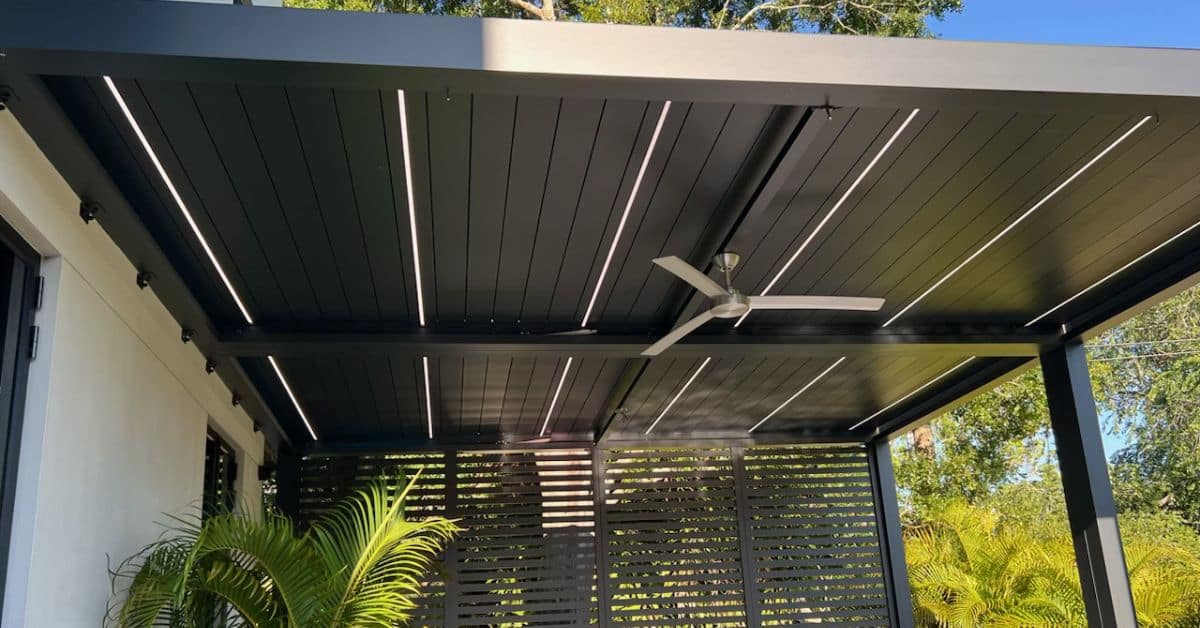 A close-up of a modern black louvered pergola ceiling with integrated white linear LED lighting strips and a sleek silver ceiling fan.