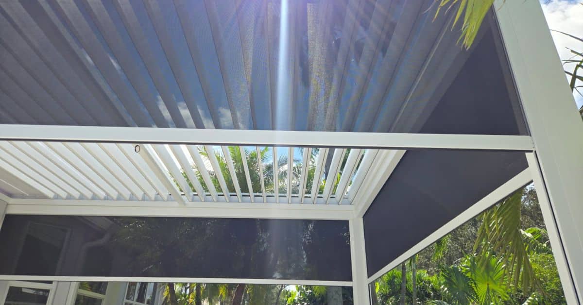 An upward view from inside a white louvered pergola featuring integrated black motorized sun screens and louvers opened to reveal a blue sky.