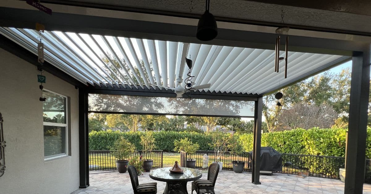 A wide view of a large dark-framed louvered pergola installed over a stone patio dining area, featuring an integrated white ceiling fan and wind chimes.