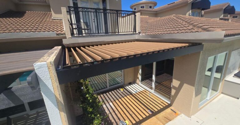 An overhead exterior view of a custom motorized louvered pergola with a dark frame and wood-grain texture louvers partially opened. The installation is built into a stucco building next to a balcony and overlooks a deck with linear shade patterns.